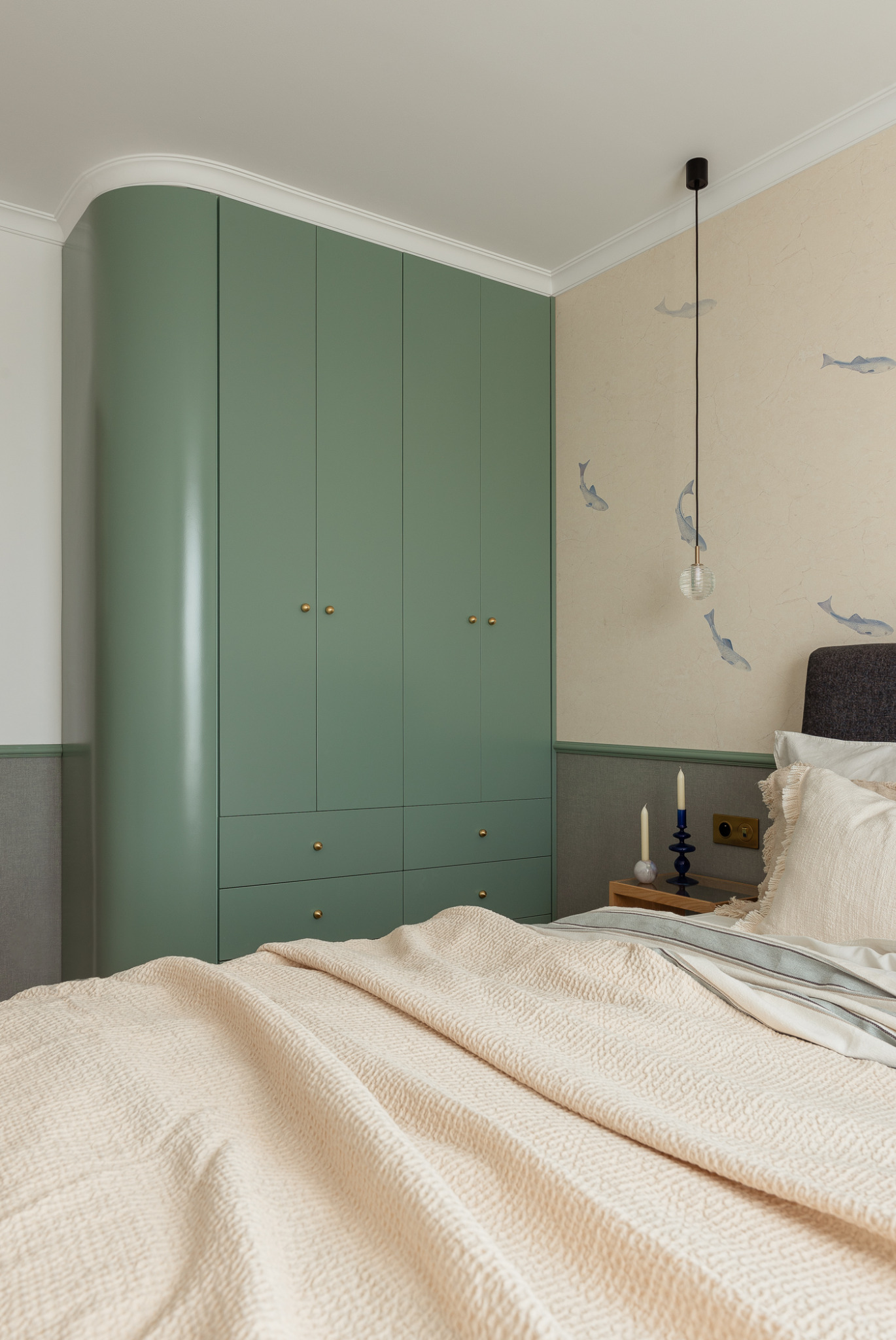 Sypialnia z pistacjową szafą z zaokrąglonym bokiem, beżową narzutą o fakturze wafelka, ścianą z motywem ryb i modernistyczną lampą wiszącą.