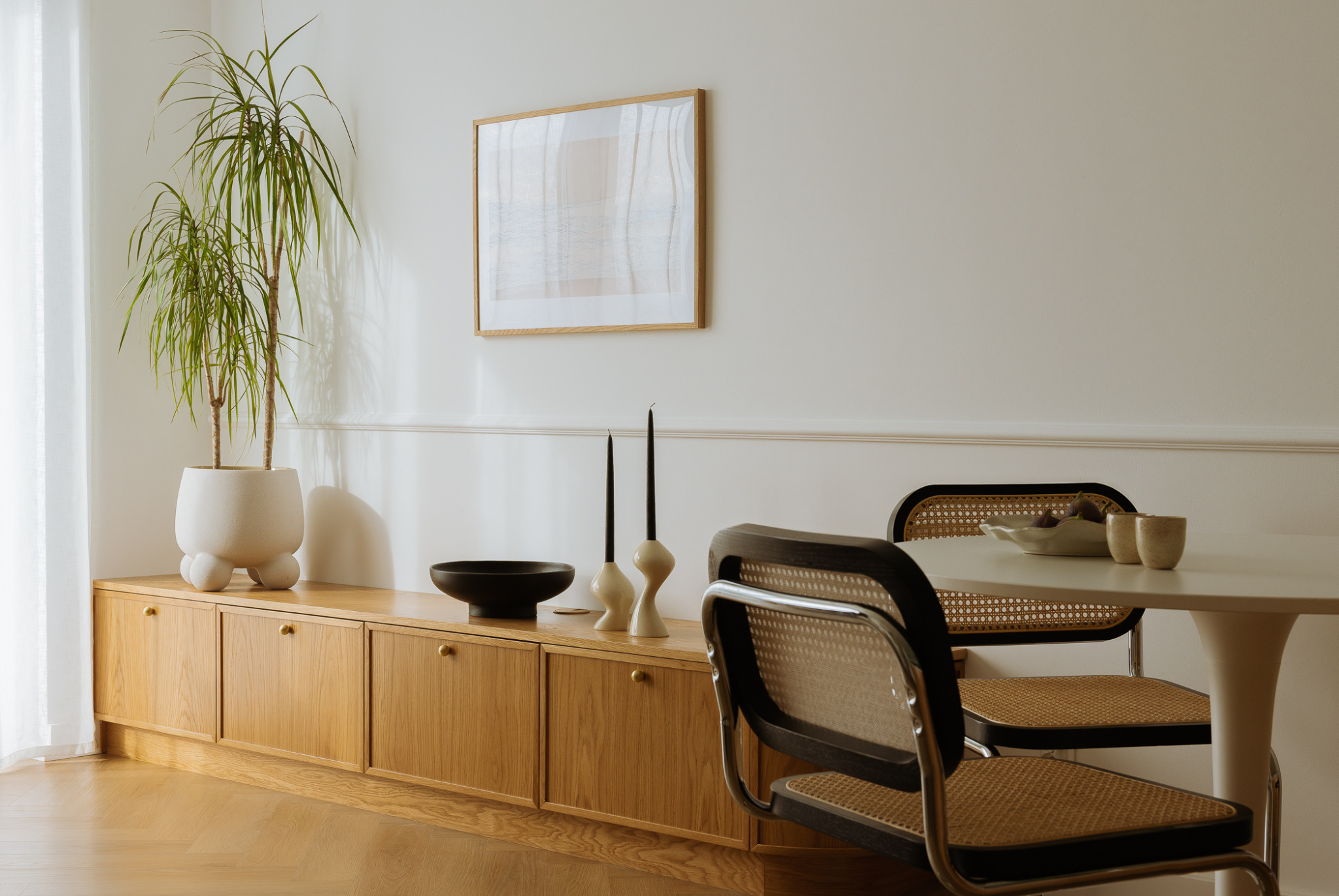 Jasne wnętrze z drewnianą komodą, na której stoi roślina w białej donicy, czarna misa i dwie świece. Okrągły biały stół z rattanowymi krzesłami. Na ścianie obraz w drewnianej ramie.