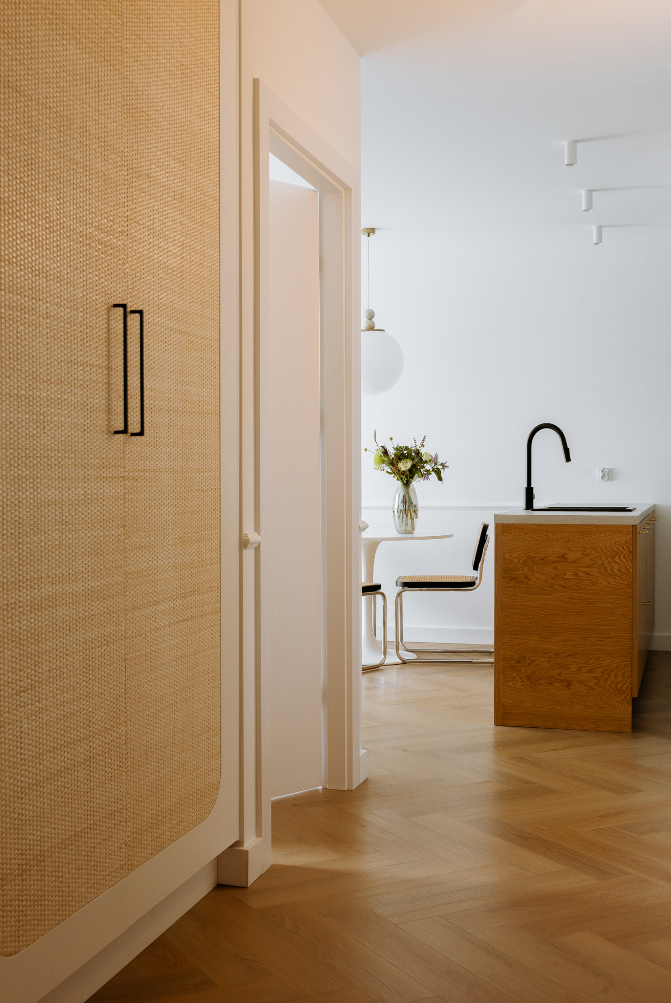 Minimalistyczne wnętrze: panel ścienny z plecionki z czarnym uchwytem, otwarte drzwi prowadzące do jadalni z okrągłym stołem i kwiatami, fragment drewnianej zabudowy kuchennej z czarną baterią...