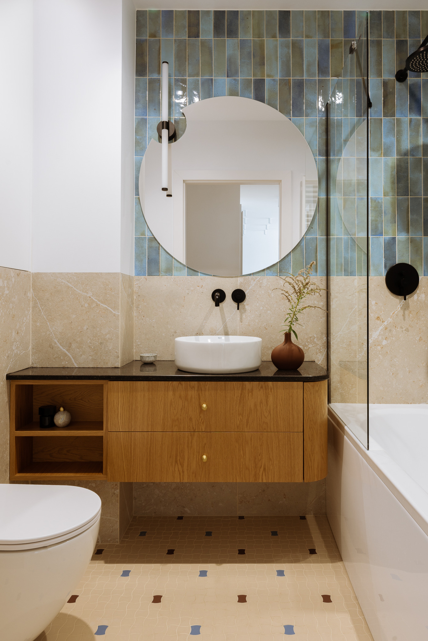 Nowoczesna łazienka z drewnianą szafką pod umywalką, okrągłym lustrem i niebieskimi płytkami na ścianie, uzupełniona brązowym wazonem z suszonymi kwiatami.