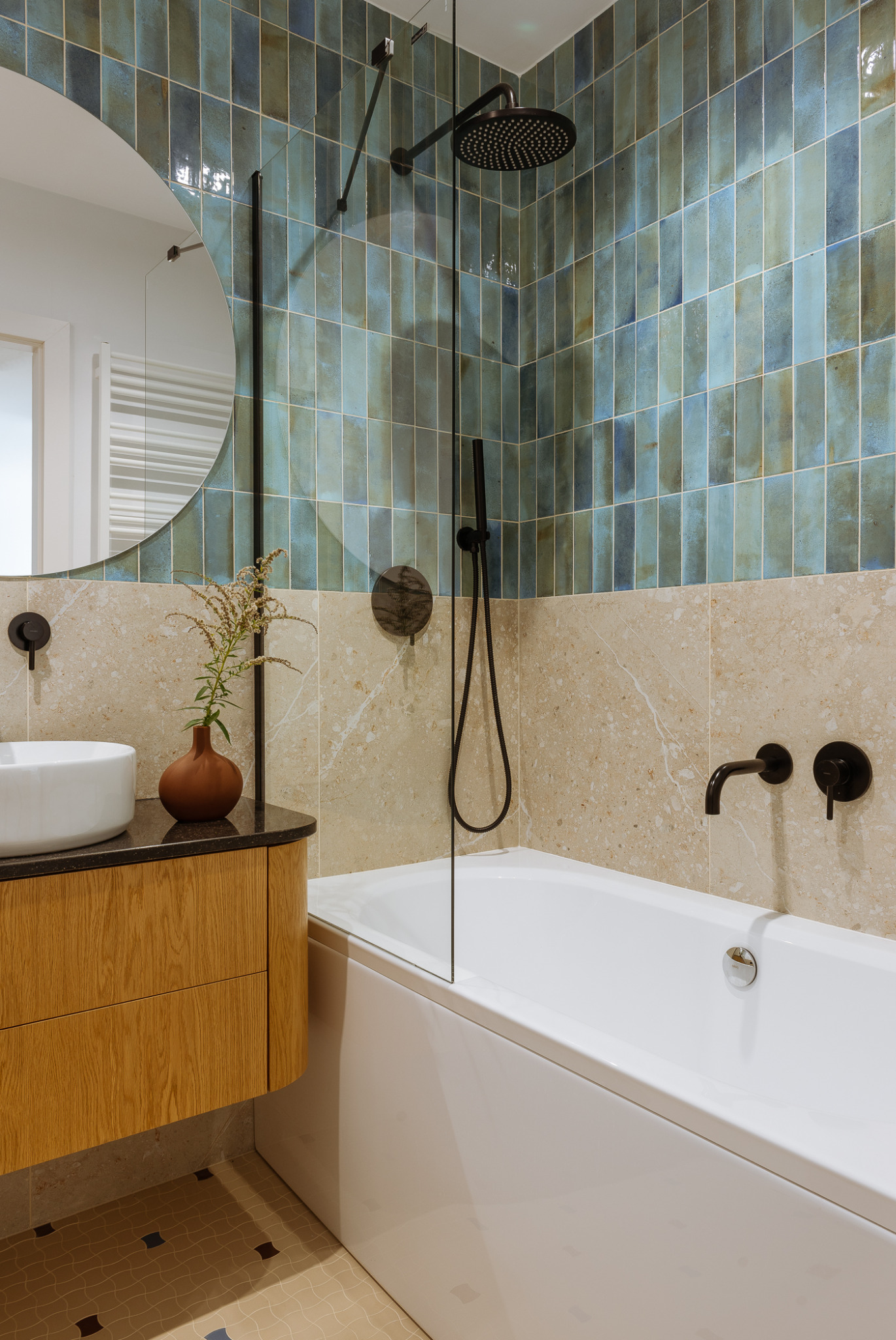 Nowoczesna łazienka z wanną obudowaną płytkami, umywalką nablatową na drewnianej szafce, okrągłym lustrem i armaturą w kolorze czarnym.