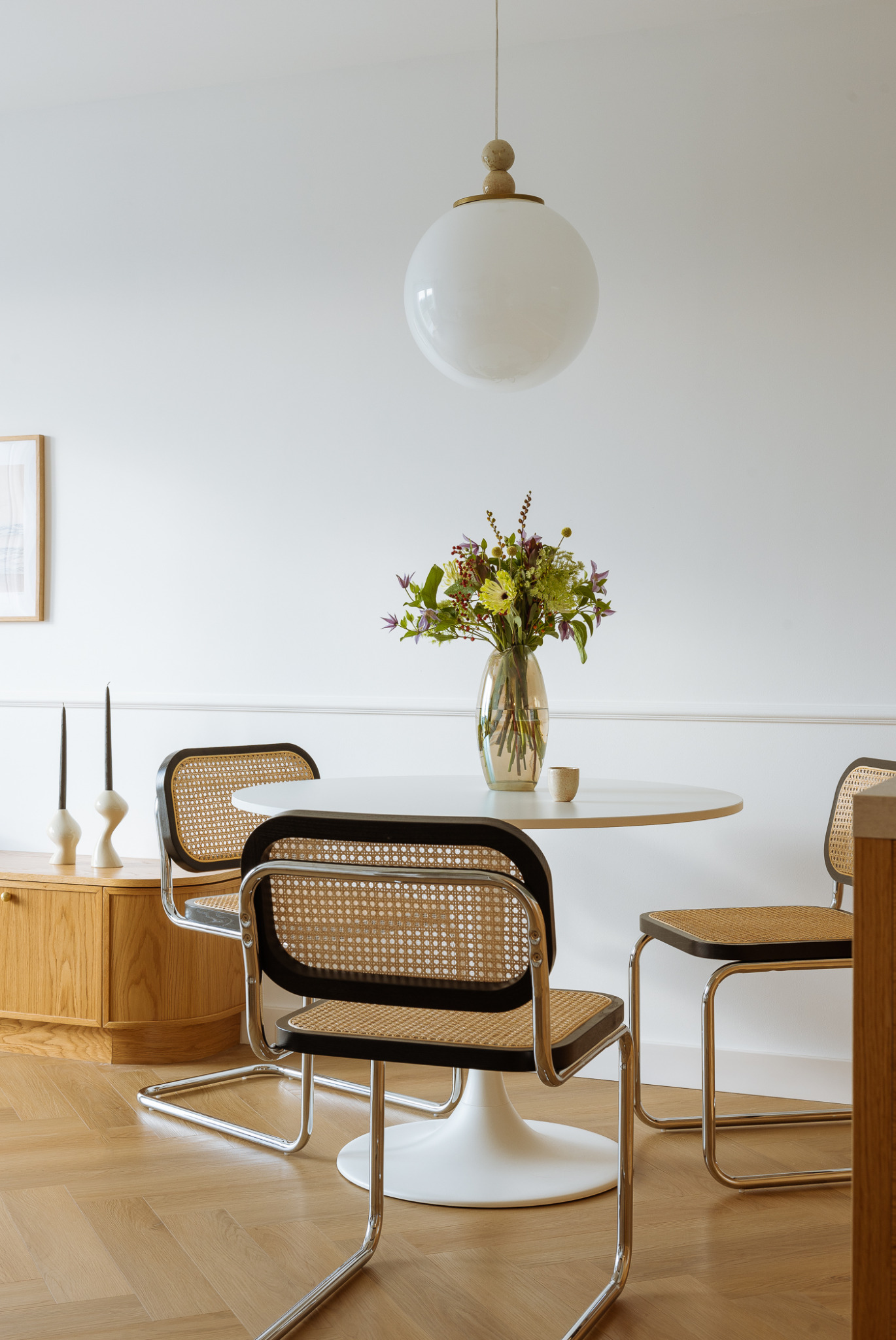 Jasne wnętrze jadalni z okrągłym, białym stołem, bukietem w szklanym wazonie, trzema krzesłami z rattanowymi siedziskami i oparciami oraz lampą kulą zwisającą z sufitu. Na komodzie widoczne wysokie...