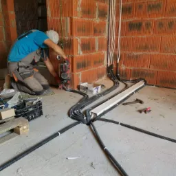 Instalator w trakcie montażu peszli z kablami w surowym wnętrzu budynku z cegły, używa wiertarki z nasadką, widoczne kable pionowo biegnące po ścianie, a peszle rozchodzą się promieniście po podłodze.