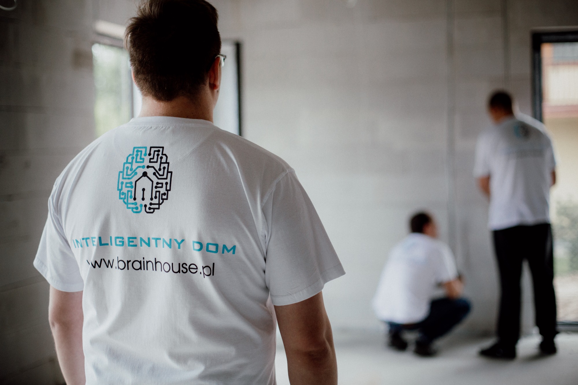 Trzech mężczyzn w białych koszulkach z logo 'Inteligentny Dom' w surowym wnętrzu, dwóch z nich pracuje, widok z tyłu na koszulkę z wyraźnym logo firmy.
