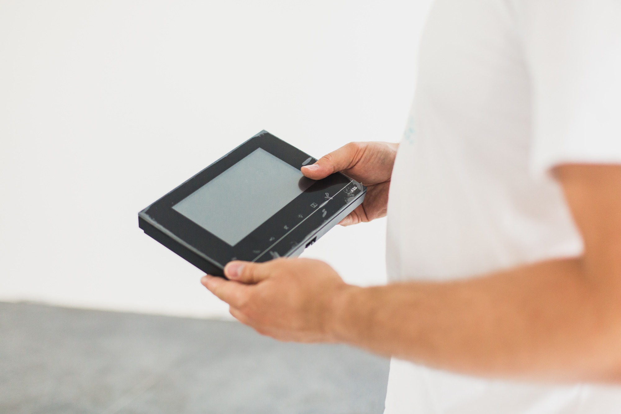 Mężczyzna w białej koszulce trzyma w dłoniach czarny panel sterowania systemem domowym z dotykowym ekranem, jeszcze w folii ochronnej.