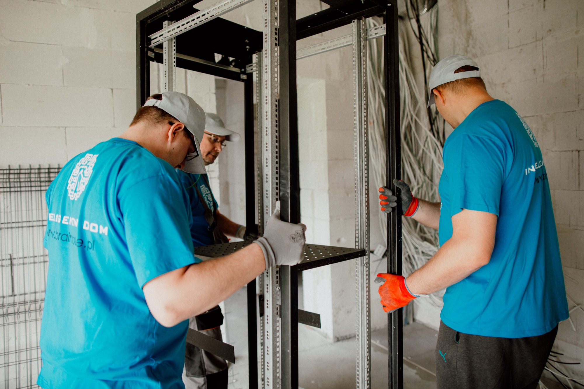 Trzech instalatorów w niebieskich koszulkach montuje metalową szafę rack w surowym pomieszczeniu z widocznymi kablami; jeden ma pomarańczowe rękawice, a na koszulkach widnieje logo i adres strony...