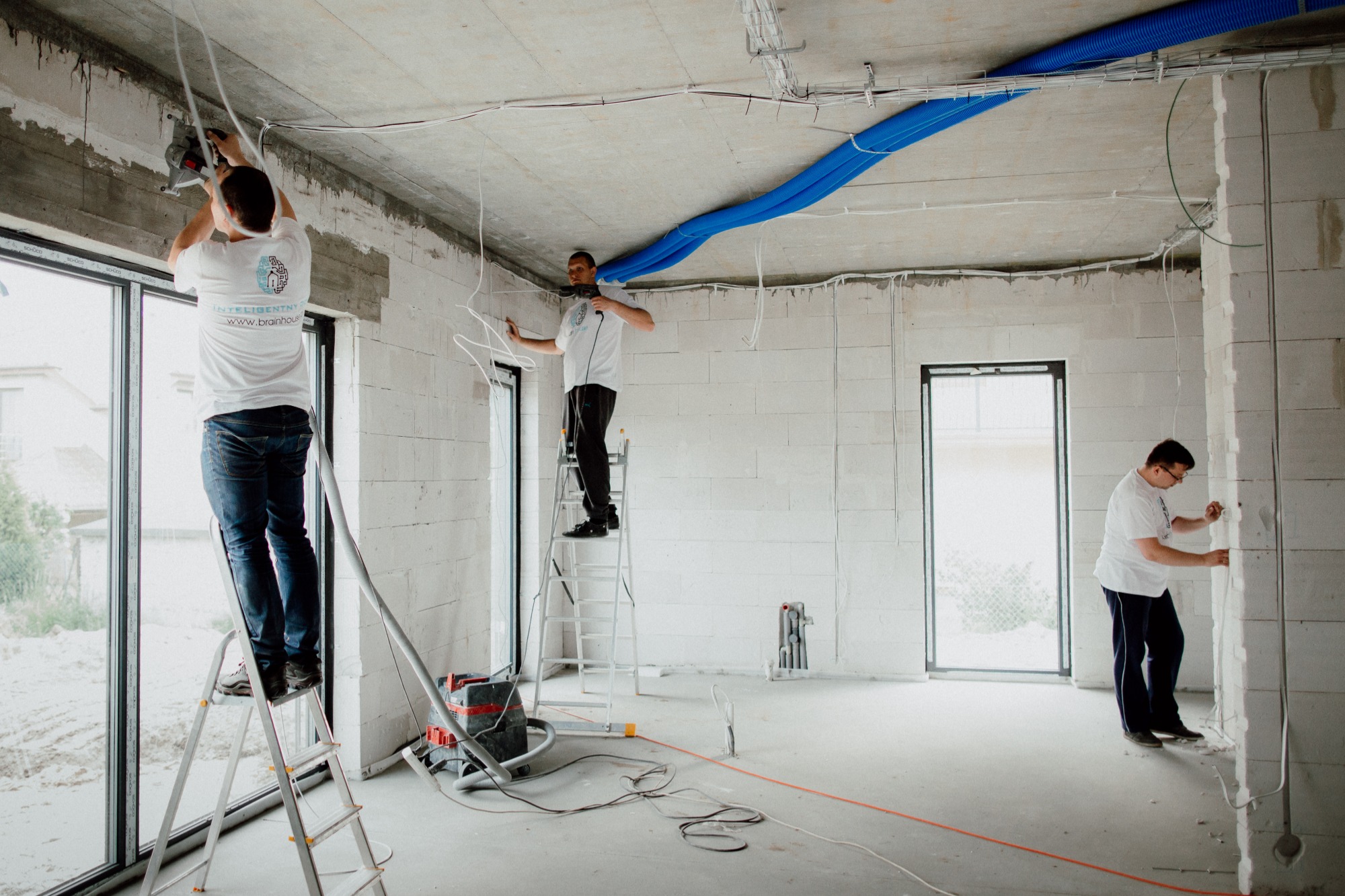 Trzech elektryków instaluje przewody w surowym stanie budynku z białych bloczków, widoczne niebieskie rury wentylacyjne pod sufitem, jeden z pracowników ma na koszulce logo firmy.