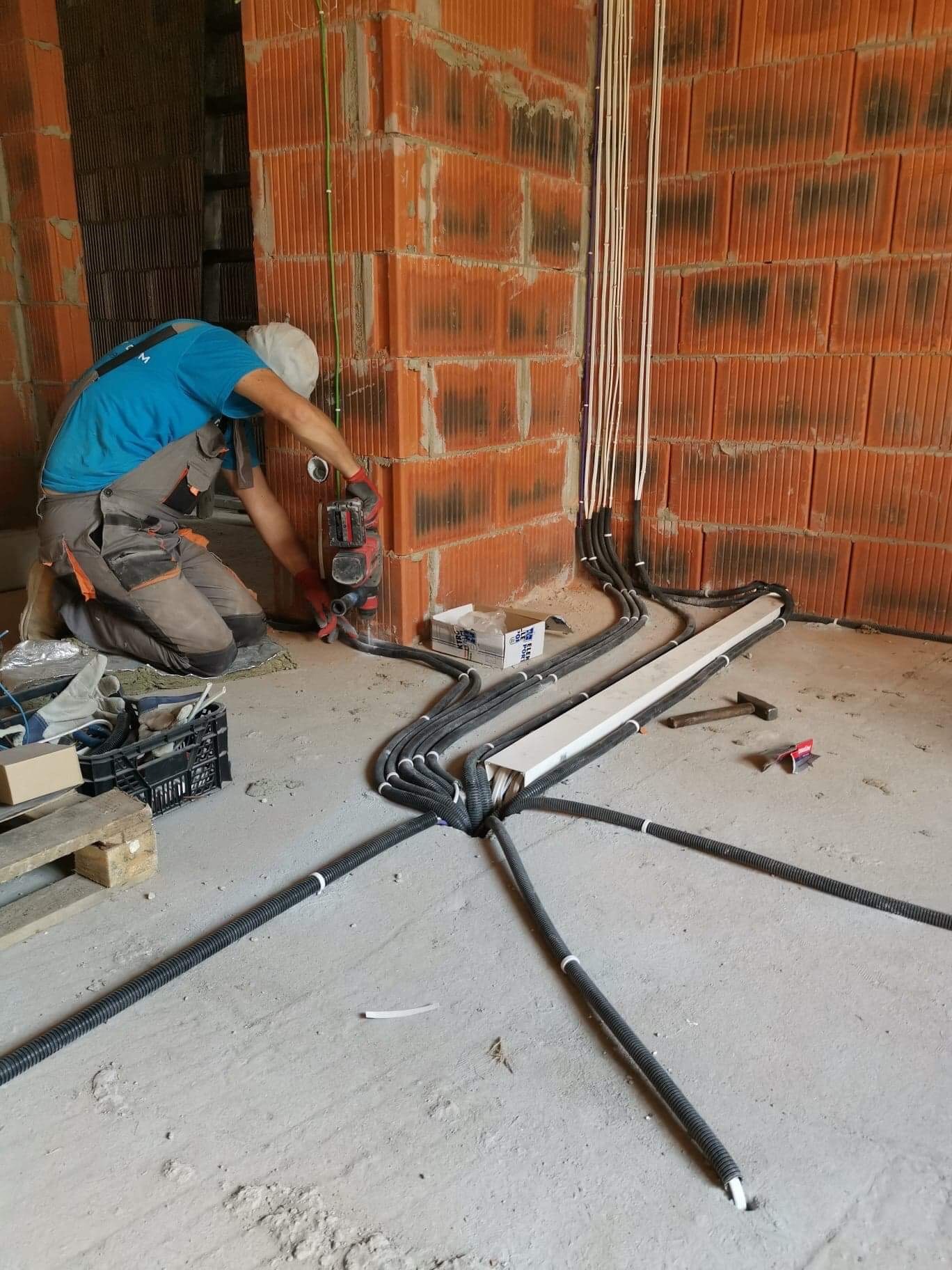 Instalator w trakcie montażu peszli z kablami w surowym wnętrzu budynku z cegły, używa wiertarki z nasadką, widoczne kable pionowo biegnące po ścianie, a peszle rozchodzą się promieniście po podłodze.