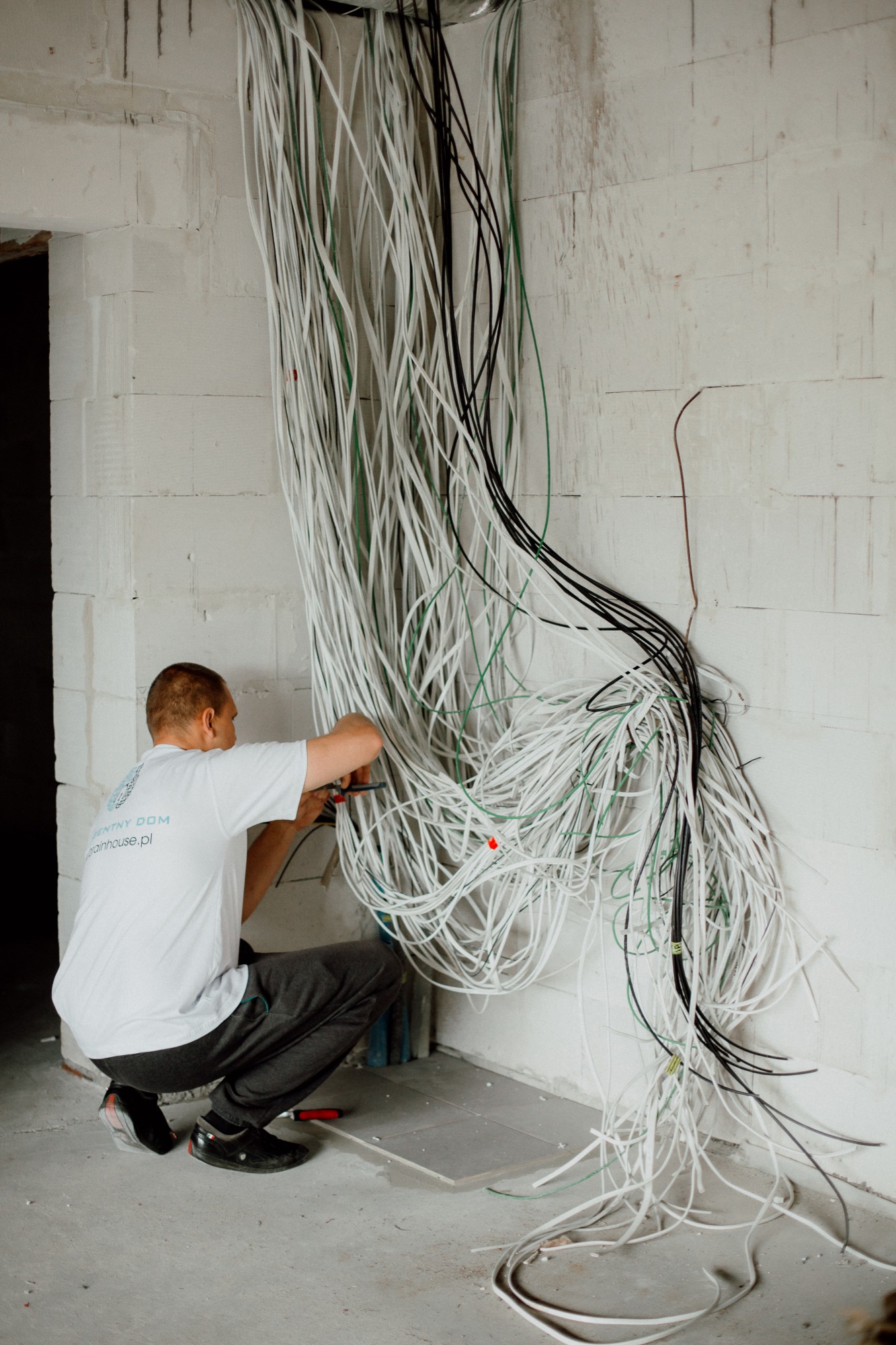 Elektryk w kucki, z narzędziami w dłoniach, montuje wiązkę przewodów elektrycznych (białe, czarne, zielone) wychodzących ze ściany z białych bloczków, na koszulce logo firmy.