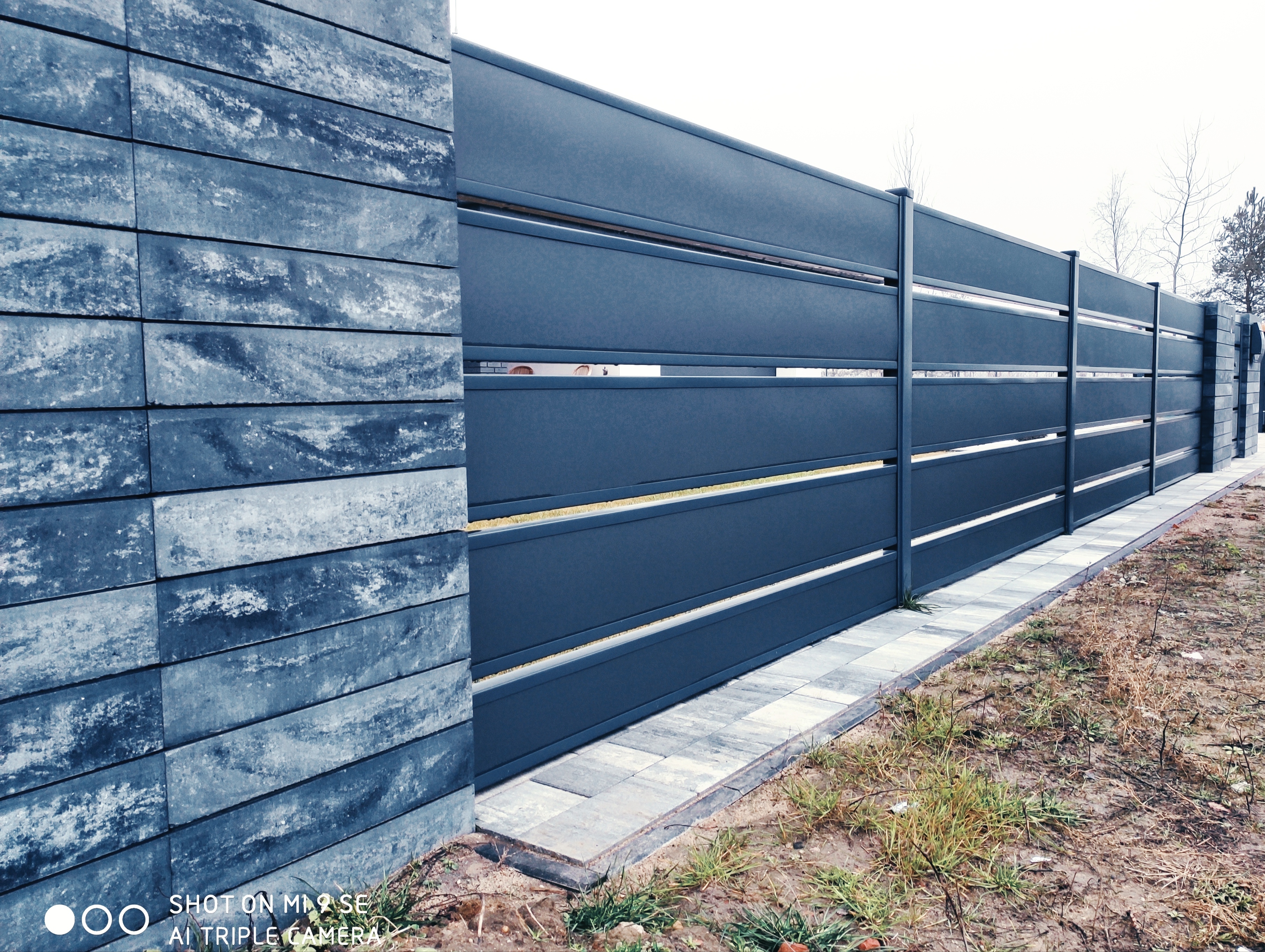 Nowoczesne, poziome przęsła w ciemnoszarym odcieniu tworzą kontrast z kamiennymi słupkami ogrodzenia w Zduńskiej Woli, a kostka brukowa podkreśla linię podziału z terenem zielonym.