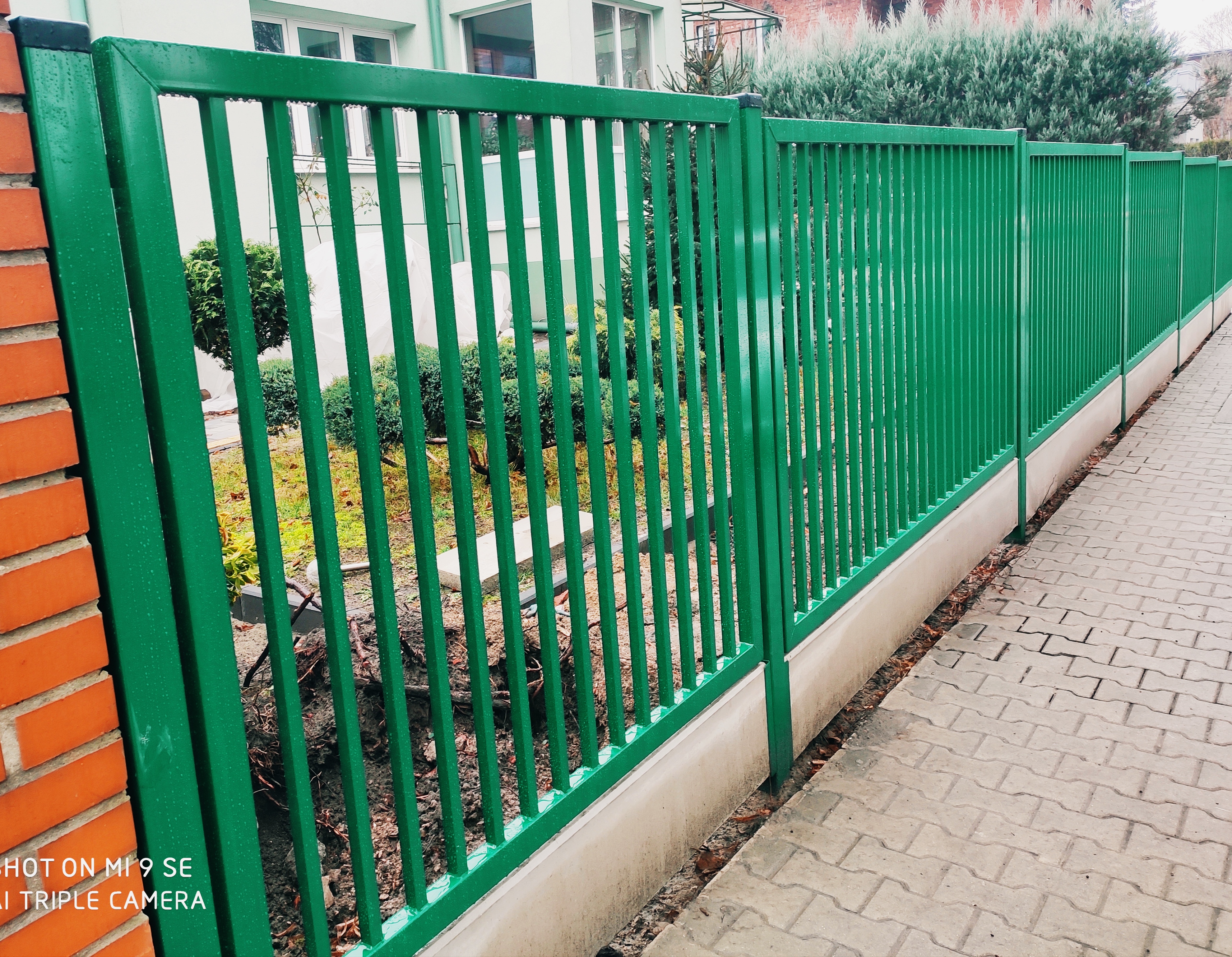Długi, zielony, metalowy płot sztachetowy z wąskimi pionowymi prętami, biegnący wzdłuż chodnika z kostki brukowej, z widoczną ceglaną ścianą po lewej stronie i zielenią w tle.