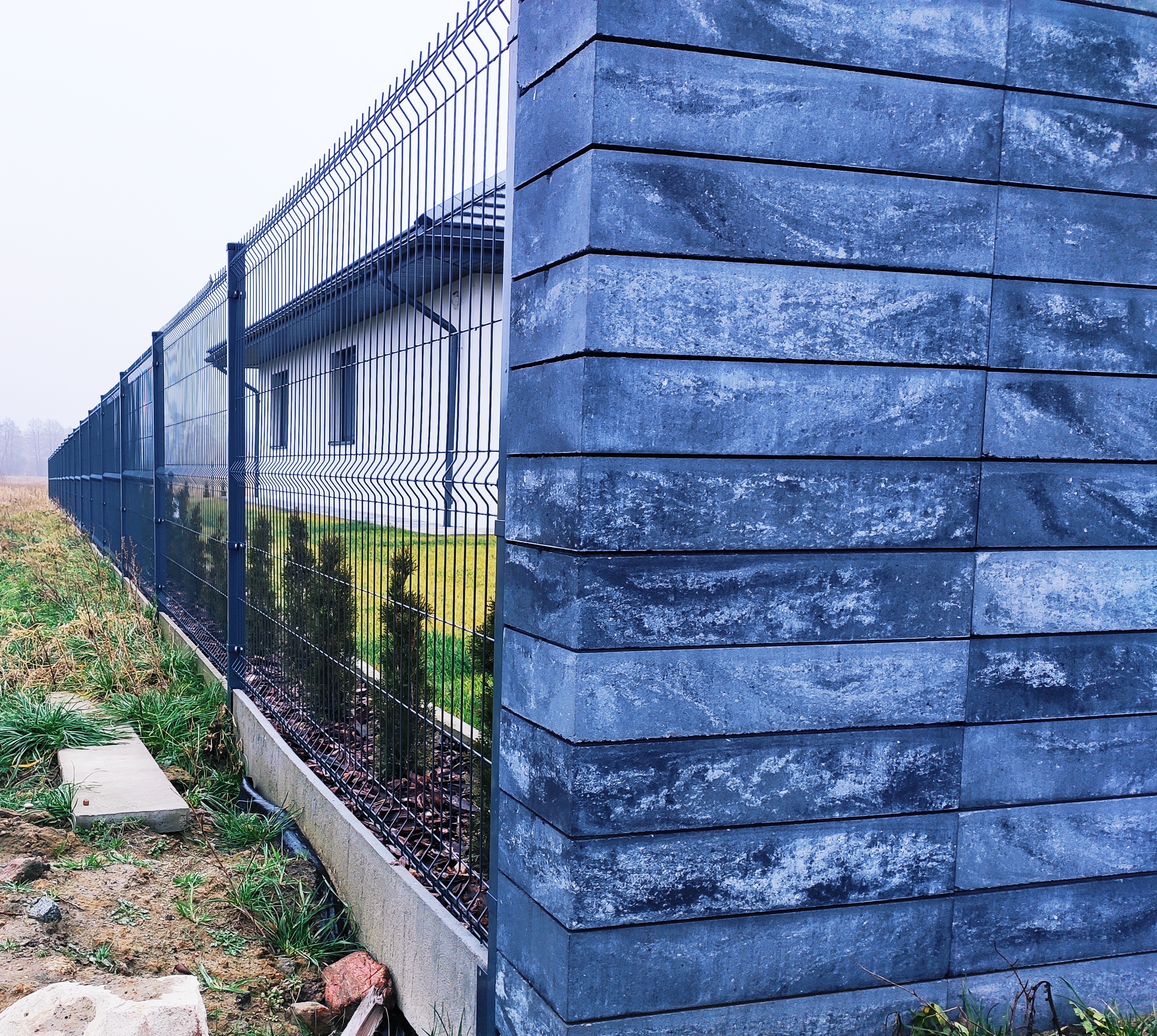 Nowoczesne ogrodzenie z paneli z podmurówką betonową i słupkami z bloczków betonowych w kolorze antracytowym, widok z boku na tle domu i zieleni.