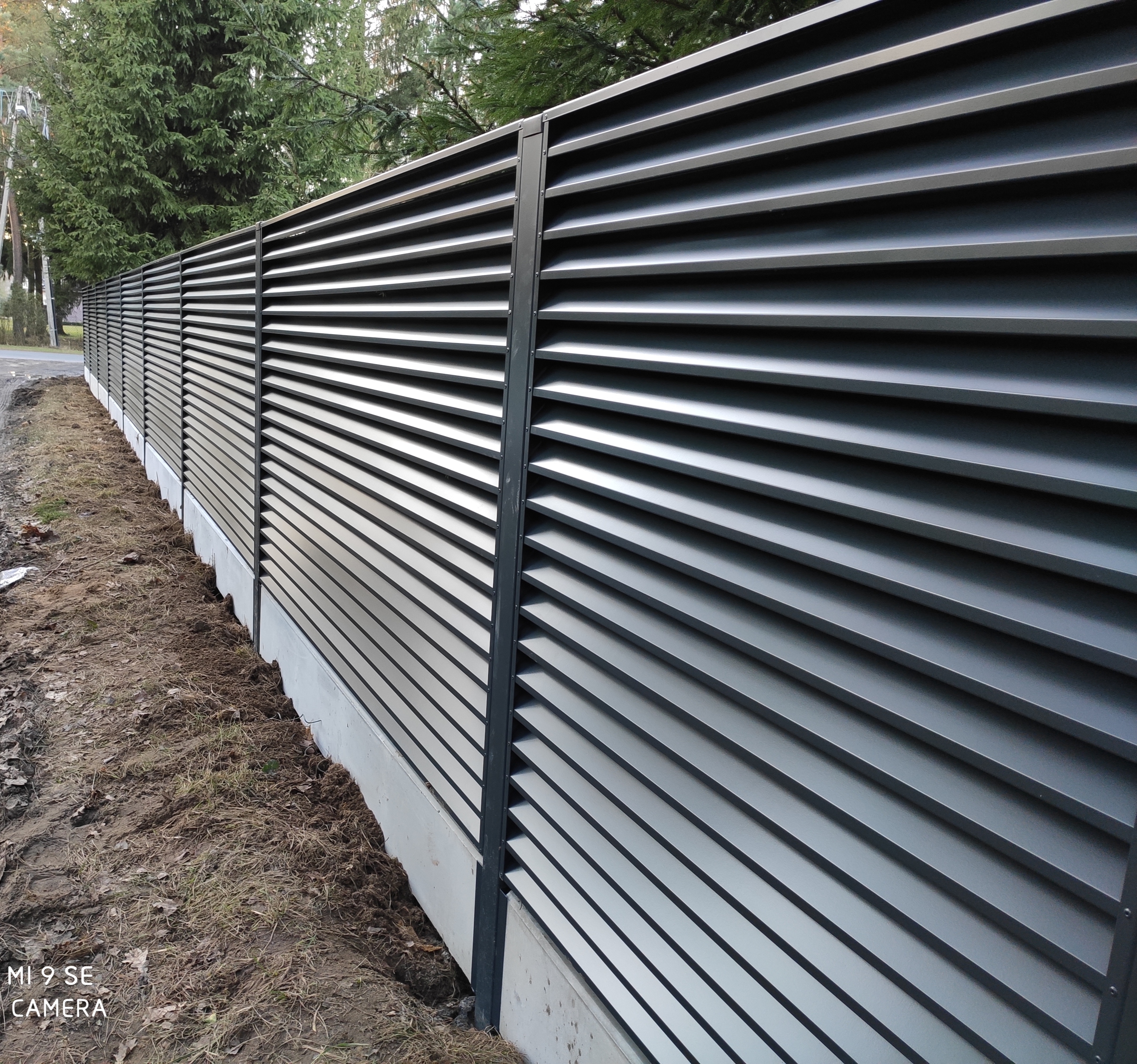 Nowoczesne, poziome przęsła w grafitowym kolorze tworzą eleganckie ogrodzenie posesji w Zduńskiej Woli. Widoczne słupki nośne i betonowa podmurówka.