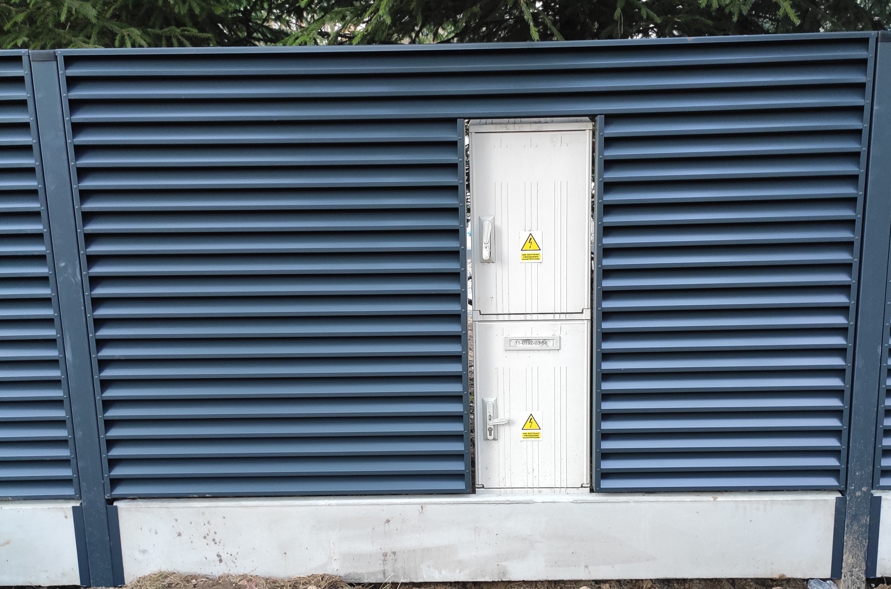 Szary panelowy płot z wbudowaną białą skrzynką elektryczną z ostrzeżeniem o wysokim napięciu.