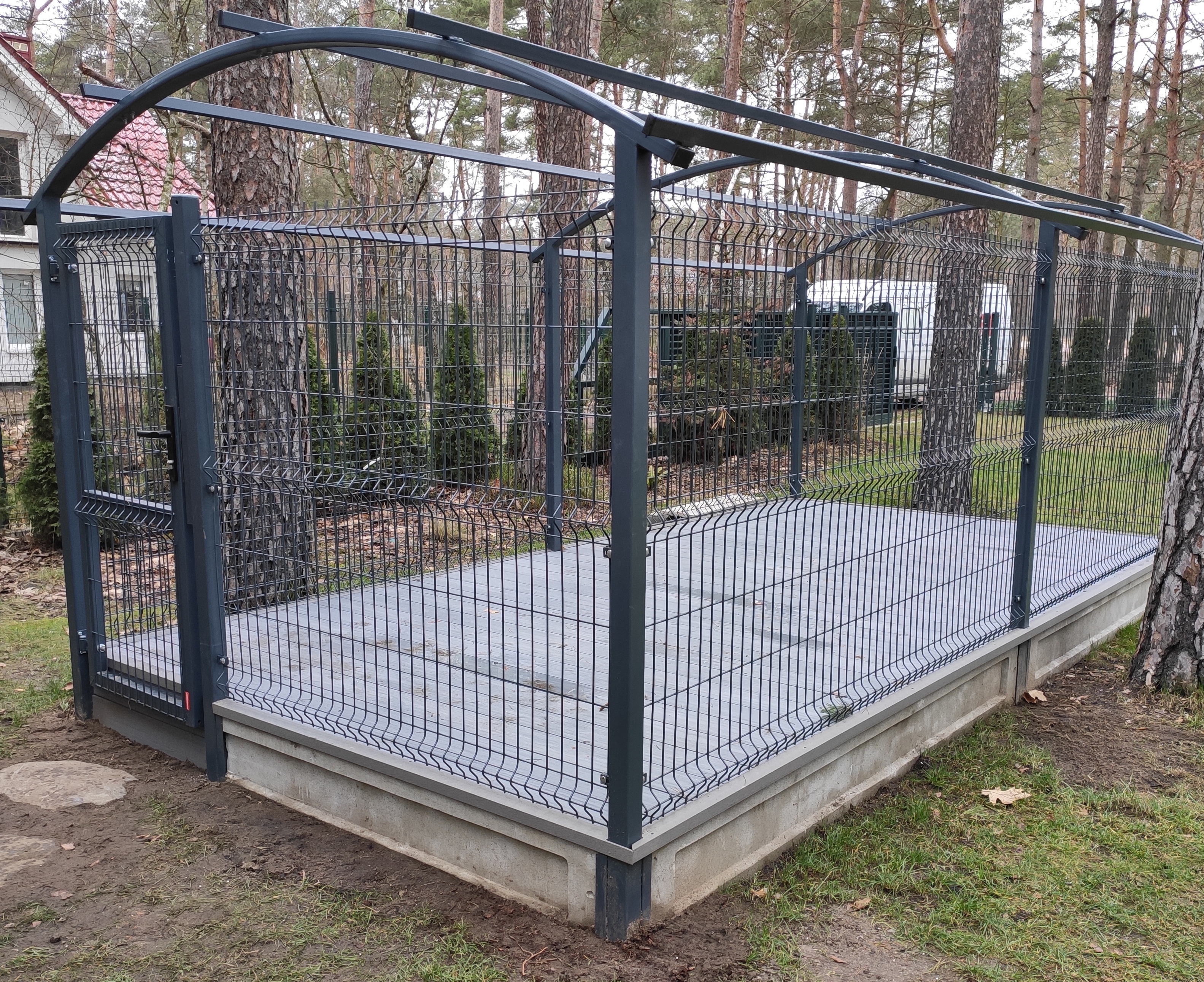 Szara, metalowa konstrukcja z siatki z bramką, otaczająca betonową platformę, w tle drzewa i dom.