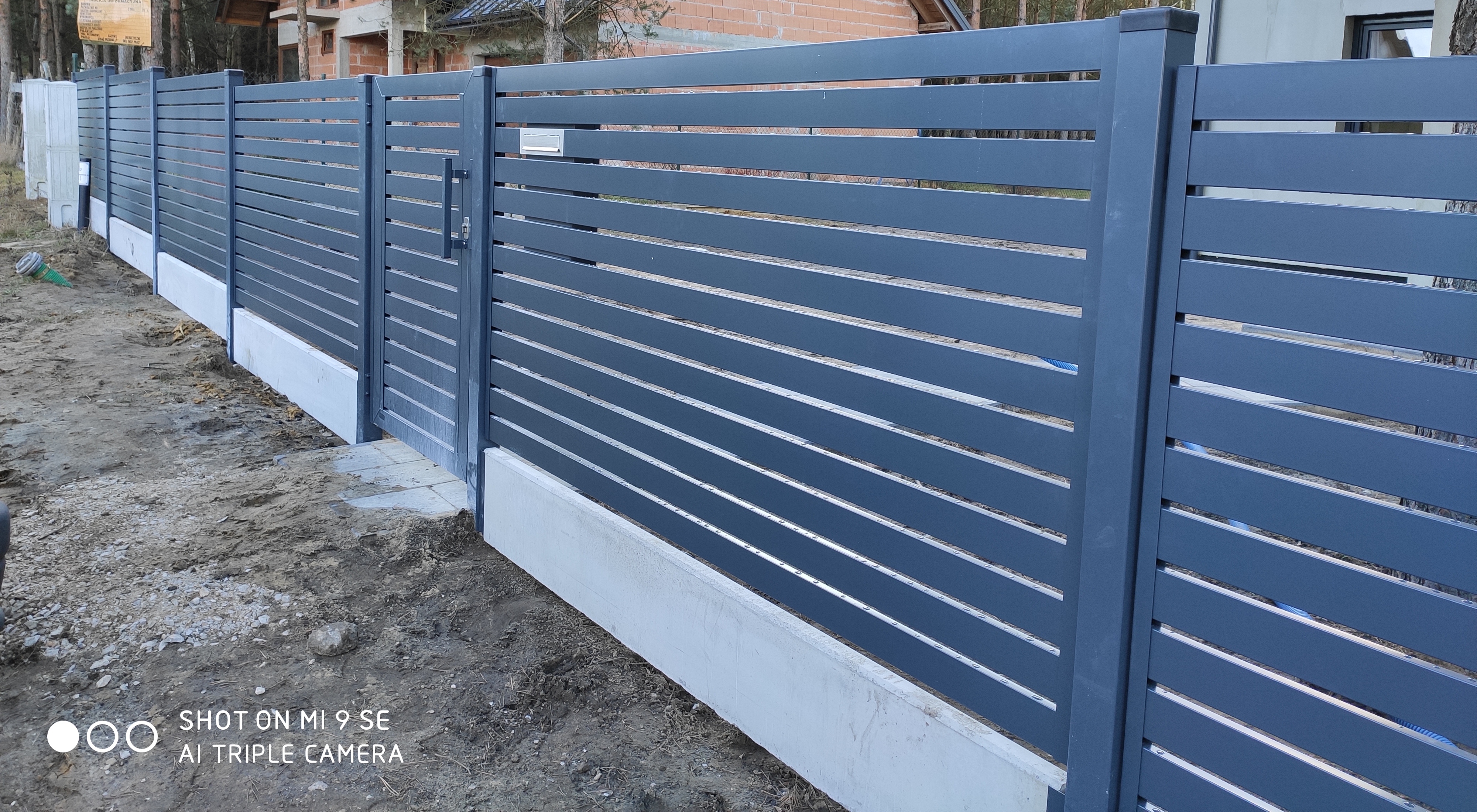 Nowoczesne, szare ogrodzenie z poziomych metalowych paneli, z bramą i betonową podmurówką, na tle drzew i budowy domu.