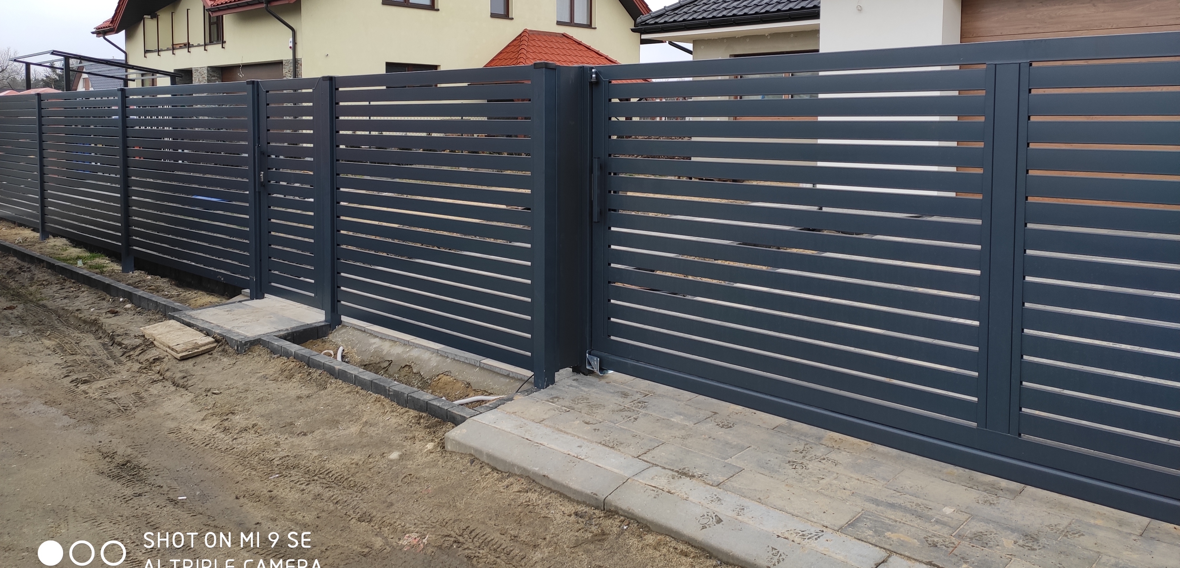 Nowoczesne, poziome, szare przęsła ogrodzenia z bramą przesuwną na posesji z kostką brukową, widok z zewnątrz.