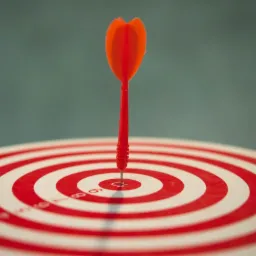 Czerwona strzałka wbita w sam środek tarczy z czerwono-białymi okręgami na tle szarej ściany, symbol precyzji i trafności.