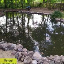 Oczko wodne otoczone dużymi, szarymi kamieniami, z odbijającymi się drzewami w tafli wody. W tle widoczny drewniany płot i taczka.