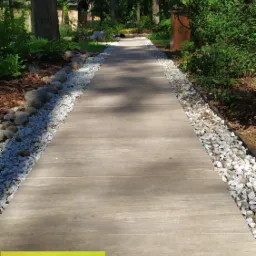 Prosta ścieżka z drewnopodobnych płyt obłożona białym grysem prowadzi przez zalesiony ogród, w tle widoczny budynek, a w rogu kadru żółta reklama usług ogrodniczych.