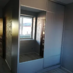 Nowoczesna szafa przesuwna z lustrem, odbijająca widok okna i fragment wnętrza pokoju z drewnianą podłogą i szarą ścianą.