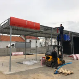 Montaż konstrukcji stalowej hali: pracownik na podnośniku widłowym montuje czerwony element dachu, w tle budynek z czerwoną dachówką, na ziemi paleta z niebieskim wężem i materiały izolacyjne.