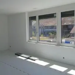 Przygotowane pomieszczenie do malowania: podłoga zabezpieczona folią malarską z taśmą, ściany świeżo pomalowane na biało, widoczne trzy okna z widokiem na budynki.