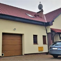 Żółty dom jednorodzinny z brązową dachówką, garażem z drewnianą bramą i szarym samochodem typu kombi na podjeździe.