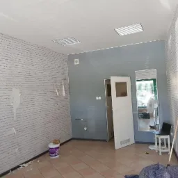 Wnętrze biura w trakcie prac przygotowawczych: ściany częściowo pokryte szarą farbą imitującą cegłę, widoczne resztki starej farby, otwarte drzwi, lustro, laptop na stołku i wiadro z farbą.
