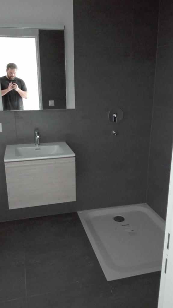 Nowoczesna łazienka z szarymi płytkami, białą umywalką z szafką z jasnego drewna i brodzikiem; w lustrze odbija się osoba robiąca zdjęcie.