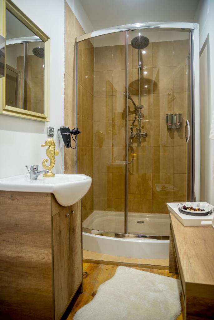 Umywalka z szafką z imitacji drewna, złoty dekoracyjny konik morski, suszarka do włosów, kabina prysznicowa z brązowymi płytkami i zestawem prysznicowym, drewniana podłoga i biały dywanik w łazience.