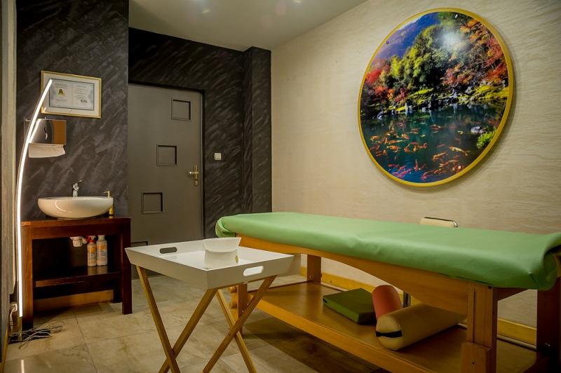 Jasne wnętrze gabinetu z łóżkiem do zabiegów, umywalką i dekoracyjnym obrazem z motywem stawu z karpiami koi na ścianie.