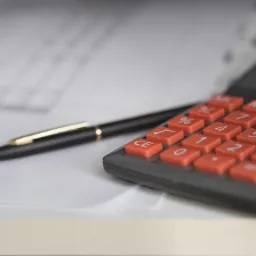 Kalkulator z pomarańczowymi przyciskami i czarny długopis na białym papierze z częściowo widocznymi danymi finansowymi w tle.