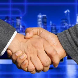 Uścisk dłoni dwóch osób w garniturach na tle rozmytego, błękitnego krajobrazu miejskiego i monitorów komputerowych.