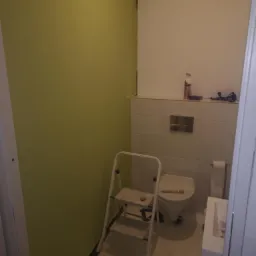 Świeżo pomalowana na zielono ściana w łazience, widoczna drabina malarska, fragment toalety i umywalki podczas remontu.