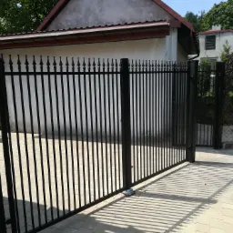 Otwarta, czarna brama metalowa z pionowymi prętami i ozdobnymi grotami, prowadząca na posesję z kostką brukową, w tle budynek z białymi ścianami i brązowym dachem oraz zieleń drzew.