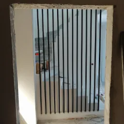 Widok przez surowy otwór drzwiowy na czarne, pionowe elementy zabezpieczające schody w trakcie remontu; widoczne niedokończone schody betonowe i przygotowania do dalszych prac wykończeniowych.