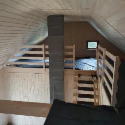 Wnętrze domu w trakcie budowy z drewnianą konstrukcją, widoczne drewniane ściany, strop i schody, komin z szarego betonu oraz czarna folia ochronna na podłodze.