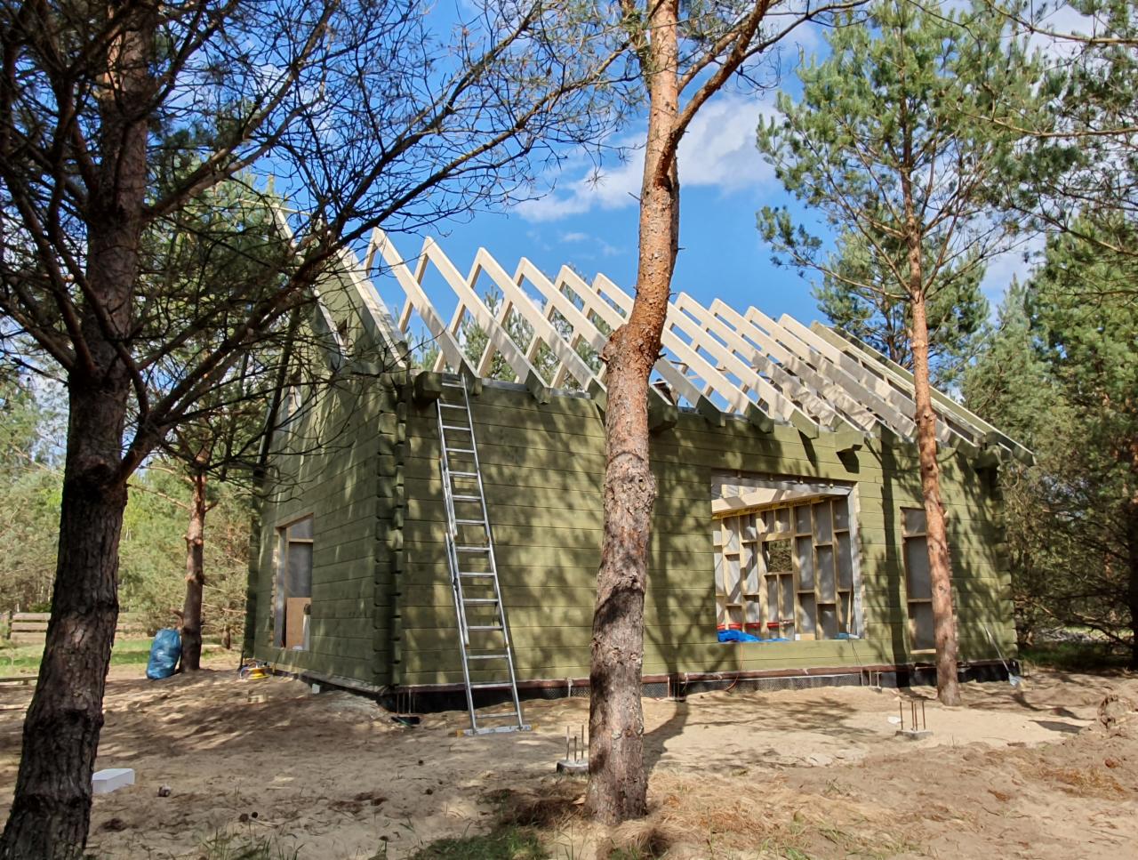 Wznoszenie domu z bali w lesie; widoczna konstrukcja dachu, ściany w kolorze butelkowej zieleni, drabina oparta o ścianę, okno z widocznym szkieletem ramy.