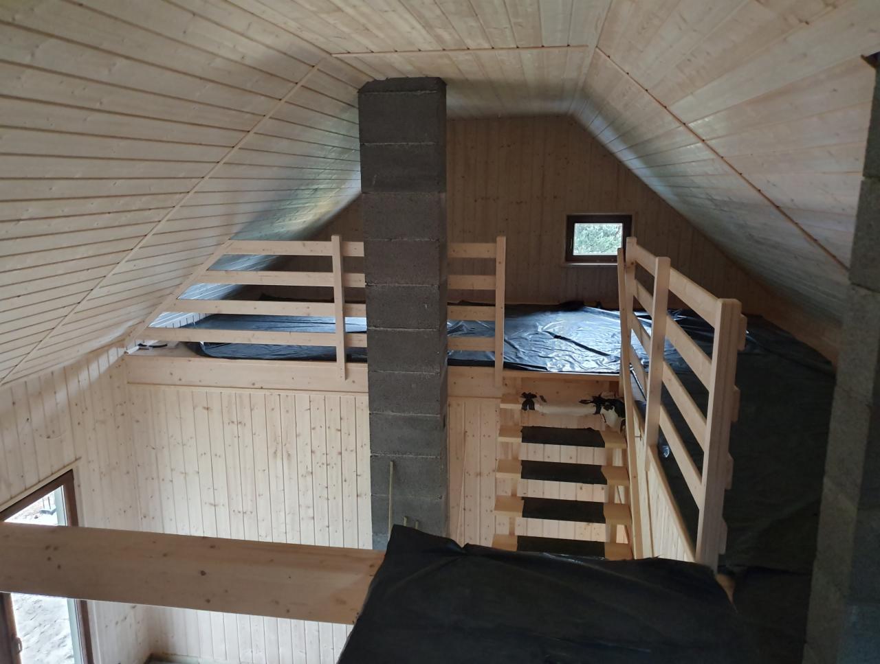 Wnętrze domu w trakcie budowy z drewnianą konstrukcją, widoczne drewniane ściany, strop i schody, komin z szarego betonu oraz czarna folia ochronna na podłodze.