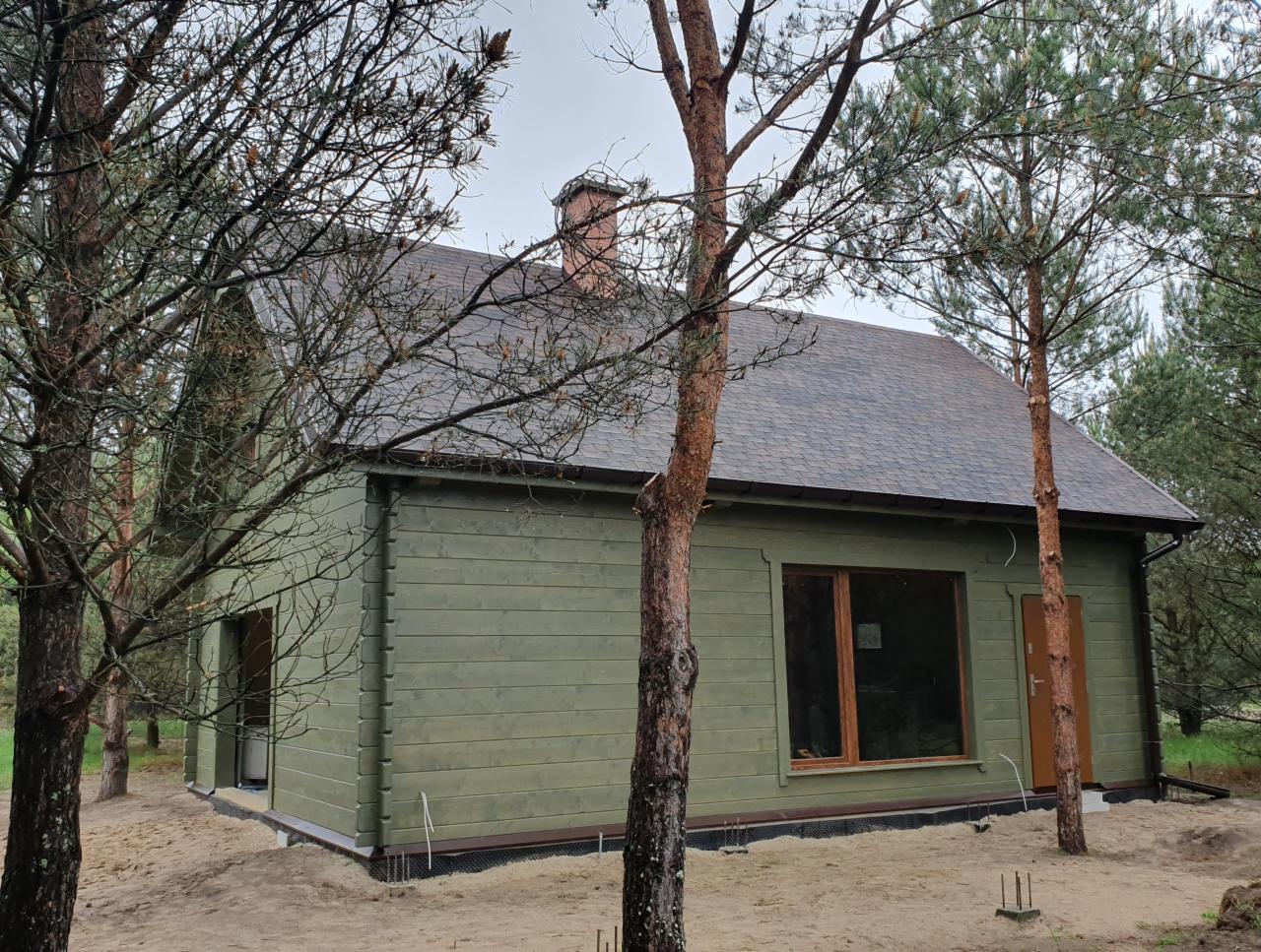Zewnętrzny widok nowo wybudowanego, parterowego domu z drewna, pomalowanego na oliwkowy kolor, z ciemnoszarym dachem bitumicznym i kominem z cegły, otoczonego młodymi sosnami.