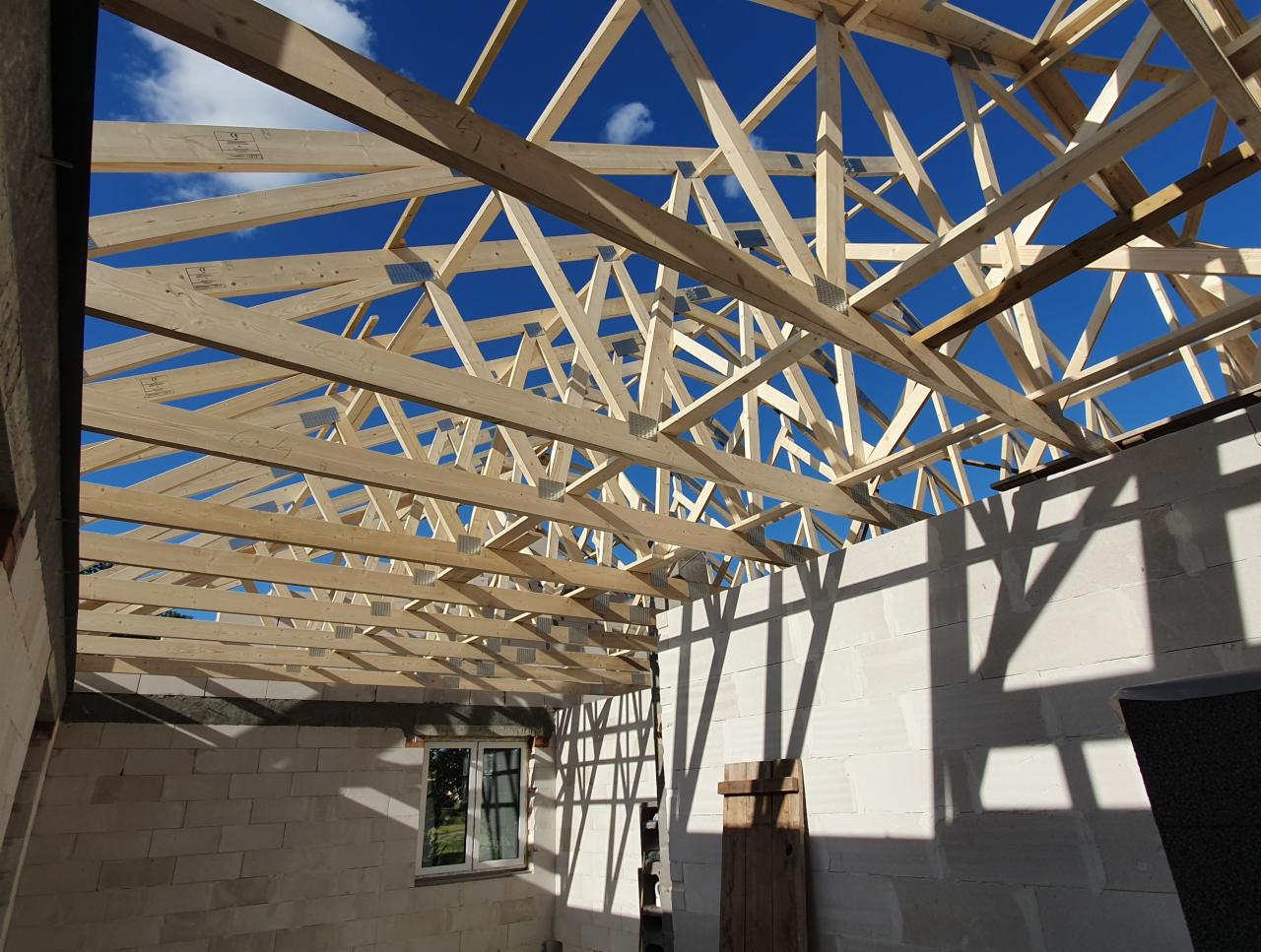 Konstrukcja dachu z drewnianych belek na tle błękitnego nieba z chmurami, widok od wewnątrz budynku z białych bloczków.