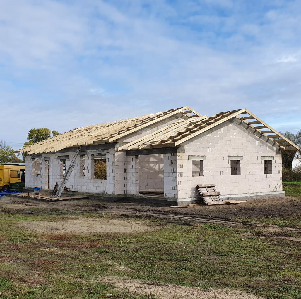 Budowa domu z betonu komórkowego w trakcie montażu więźby dachowej, widoczna drabina oparta o ścianę, żółty samochód dostawczy w tle.