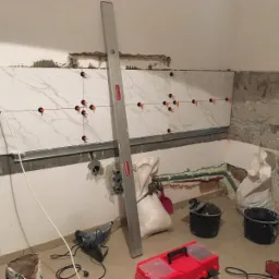 Ściana w trakcie układania płytek imitujących marmur, z użyciem poziomicy i systemu poziomowania; widoczne narzędzia i materiały budowlane w nieuporządkowanym otoczeniu.
