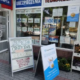 Fasada budynku z szyldami firm ubezpieczeniowych i tłumaczeń, tablice reklamowe na chodniku oferujące niskie opłaty i wysyłkę paczek.