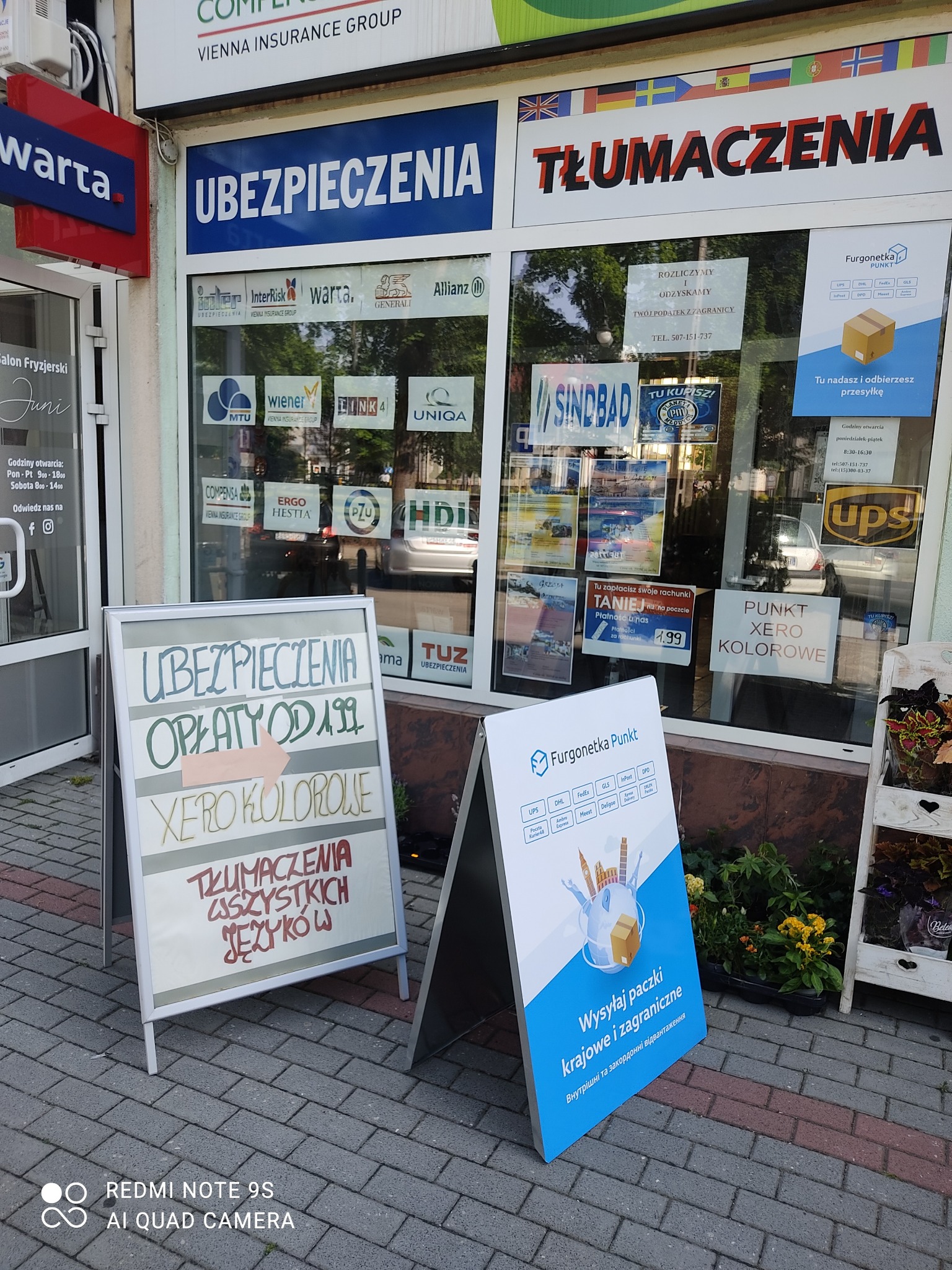 Fasada budynku z szyldami firm ubezpieczeniowych i tłumaczeń, tablice reklamowe na chodniku oferujące niskie opłaty i wysyłkę paczek.