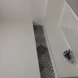 Biała, nowoczesna łazienka z błyszczącymi, białymi płytkami ściennymi i podłogą z dekoracyjnymi, czarno-białymi płytkami wzorzystymi, widoczna biała wanna z chromowaną baterią.