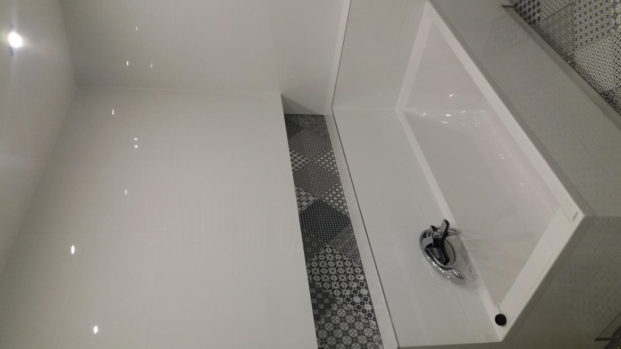 Biała, nowoczesna łazienka z błyszczącymi, białymi płytkami ściennymi i podłogą z dekoracyjnymi, czarno-białymi płytkami wzorzystymi, widoczna biała wanna z chromowaną baterią.