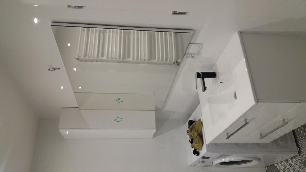 Nowoczesna, biała łazienka z umywalką nablatową z czarną armaturą, szafką pod umywalką, pralką i grzejnikiem drabinkowym. Widoczne białe płytki na ścianach i szara mozaika na podłodze.