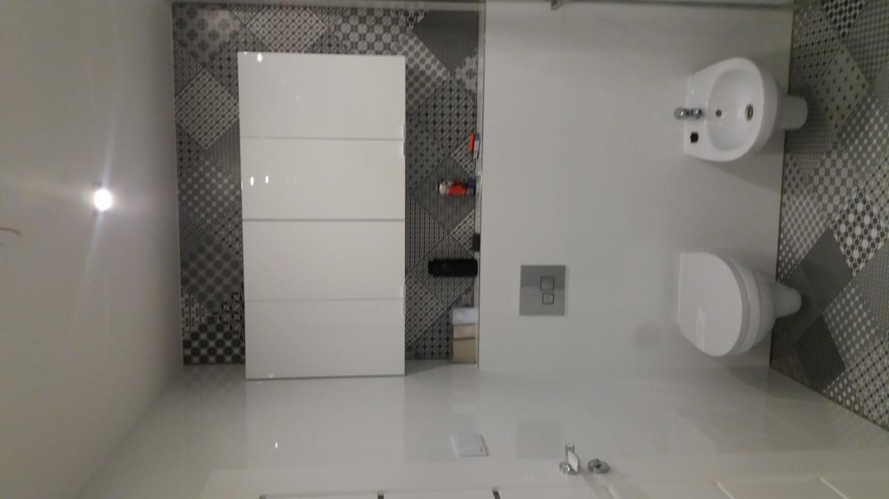 Nowoczesna łazienka z białą armaturą i geometrycznymi płytkami w odcieniach szarości, widok na umywalkę typu bidet i toaletę, z białymi szafkami po lewej stronie.